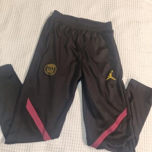 Svarta träningsbyxor PSG - Svarta träningsbyxor med PSG-logga och guldfärgad Jumpman. Byxorna har elastisk midja och rosa detaljer längs benen. 