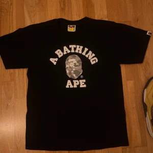 Svart t-shirt från A Bathing Ape - Säljer en svart t-shirt från A Bathing Ape med deras ikoniska logga i camo-tryck på bröstet. T-shirten har korta ärmar och en klassisk rund halsringning. Perfekt för streetwear-entusiaster!