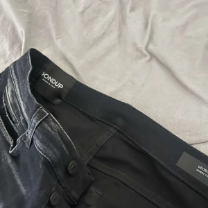 Svarta jeans från Dondup - Snygga svarta jeans från Dondup med slitna detaljer och coola färgstänk. De har en slim passform och är perfekta för en trendig look. Jeansen har knappar och klassiska fem fickor.