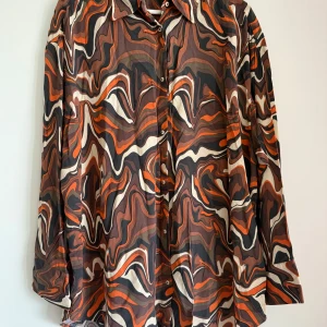 Mönstrad skjorta från Kappahl - Säljer en färgglad skjorta från Kappahl med ett abstrakt mönster i brunt, orange och beige. Skjortan har långa ärmar och knappar framtill. Perfekt för dig som vill sticka ut med en unik stil!
