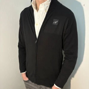 Svart kofta från Ted Bernhardtz - Snygg svart kofta från Ted Bernhardtz med dragkedja och ribbad krage. Perfekt för en stilren look. Den har en diskret logga på bröstet och är tillverkad i ett mjukt material. Plagget sitter normalt för sin storlek. Modell på bild är 182cm/66kg.  Hör gärna av dig vid fler frågor. Mvh CAVI.