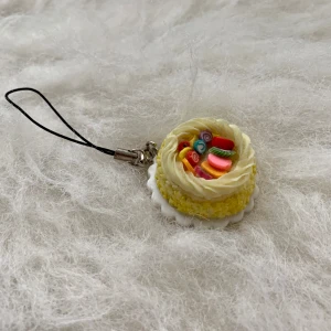 Gul tårtformad mobilaccessoar - Charmig mobilaccessoar i form av en gul tårta med färgglada detaljer på toppen. Perfekt för att ge din mobil en söt och unik touch. Fästes enkelt med en svart rem.