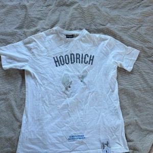 Vit t-shirt från Hoodrich - Säljer en vit t-shirt från Hoodrich med tryck både fram och bak. Framsidan har texten 'HOODRICH' i grått och baksidan har ett blått tryck med texten 'From Nothing To Something'. Perfekt för en avslappnad stil.