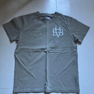Beige t-shirt från Bvaldi - Säljer en stilren beige t-shirt från Bvaldi med ett broderat vitt emblem på bröstet. T-shirten har en klassisk rund hals och korta ärmar. Perfekt för en avslappnad look.