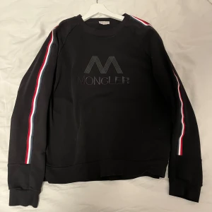 Svart sweatshirt från Moncler - Snygg svart sweatshirt från Moncler med deras logga på bröstet och ett märke på baksidan. Tröjan har långa ärmar med ränder i rött, vitt och blått längs sidorna. Perfekt för en stilren och sportig look. Pris sänks vid snabb affär