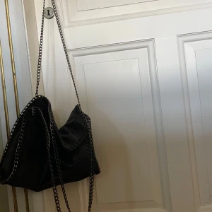 Svart axelväska från Stella McCartney - Snygg svart axelväska från Stella McCartney med kedjedetaljer. Väskan har en stilren design och är perfekt för att bära dina dagliga nödvändigheter. Insidan är fodrad med ett mjukt material i en kontrasterande färg.