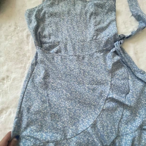 Blå blommig omlottklänning - Charmig blå omlottklänning med ett fint blommönster. Klänningen har smala axelband och knytning i midjan för en justerbar passform. Perfekt för en somrig look! Aldrig använd💙