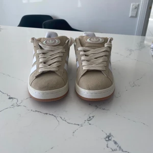 Beige Adidas sneakers - Snygga beige sneakers från Adidas med vita detaljer och klassiska ränder. Skorna har en gummisula i ljusbrun färg och snörning framtill. Perfekta för en stilren och avslappnad look.