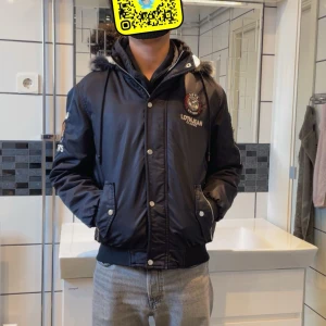 Svart bomberjacka med pälsfodrad huva - Snygg svart bomberjacka från Lothian med pälsfodrad huva och broderade detaljer. Jackan har dragkedja och knappar framtill samt ribbade muddar. Perfekt för kyliga dagar med sin varma fleecefoder.