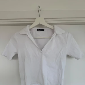 Vit ribbad topp från Zara - Säljer en stilren vit ribbad topp från Zara med korta ärmar och en klassisk krage. Toppen har en v-ringad design samt något cropped