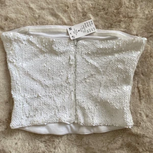 Vit topp från Gina Tricot - Säljer en snygg vit topp från Gina Tricot, täckt med glittrande paljetter. Perfekt för festliga tillfällen. Toppen är axelbandslös och har en tight passform. 