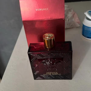 Versace Eros Flame Eau de Parfum - Säljer en elegant Versace Eros Flame Eau de Parfum. Flaskan är mörkröd med ett gyllene lock och har det ikoniska Versace-emblemet. Förpackningen är i en matchande röd färg med guldtext. Perfekt för den som vill ha en lyxig doftupplevelse.