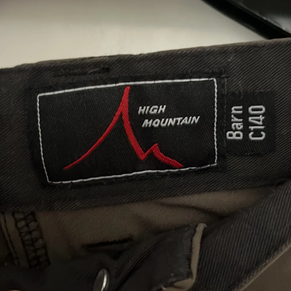 Säljer ett par funktionella byxor från High Mountain i brunt och svart. Byxorna har en praktisk design med knappar och dragkedja. Perfekta för utomhusaktiviteter och äventyr.. Farkut & Housut.