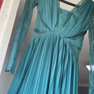 Turkos långklänning med spetsdetaljer - Elegant turkos långklänning med plisserad kjol och spetsdetaljer. Klänningen har långa ärmar och en v-ringad design. Perfekt för speciella tillfällen.