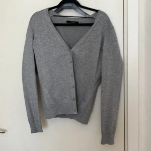 Grå kofta från Vero Moda - Säljer en stilren grå kofta från Vero Moda. Den har en v-ringad design med knappar framtill och långa ärmar. Perfekt för lager-på-lager under kyligare dagar.