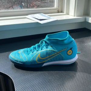 Turkosa fotbollsskor från Nike - Snygga turkosa fotbollsskor från Nike med gula detaljer och snörning. Skorna har en strumpliknande passform och är designade för inomhusbruk. Perfekta för den som vill ha stil och funktion på planen.    Använda en säsong..      om ni har någon fråga så är det bara att höra av sig.