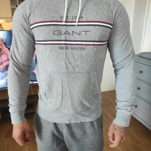 Grå hoodie från GANT - Säljer en stilren grå hoodie från GANT med texten 'NEW HAVEN' och 'EST. 1949' tryckt på bröstet. Tröjan har en känguruficka och justerbar huva med snören. Perfekt för en avslappnad look.