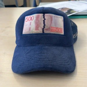 All Cap one ceps limited edition  - Snygg blå keps med ett unikt sedelmotiv på framsidan. Kepsen har ett nätmaterial på baksidan för extra ventilation och justerbar rem för perfekt passform. Perfekt för en avslappnad stil.