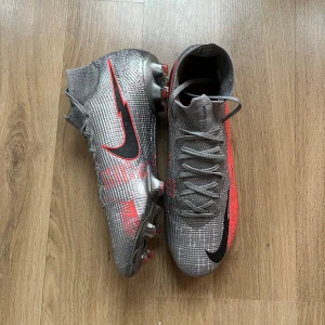 Nike fotbollsskor - Snygga fotbollsskor från Nike i grått med röda och svarta detaljer. Skorna har en modern design med snörning och en rund tå. Perfekta för fotbollsplanen med sina greppvänliga dobbar.