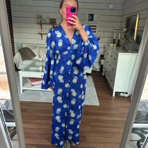 Blå blommig långklänning från Adoore - Säljer en vacker blå långklänning med vita blommor i den populära postitano-modellen från Adoore. Klänningen har långa, vida ärmar och en v-ringad design med knappar framtill. Perfekt för sommarens festligheter! Den saknar en knapp längst ner och har fått små revor i tyget men inget som påverkar utseendet, se bilder!