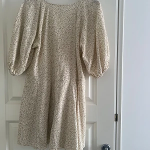 Beige blommig klänning från H&M - Charmig beige klänning från H&M med ett subtilt blommönster. Klänningen har en v-ringad design och puffiga trekvartsärmar som ger en romantisk touch. Perfekt för vårens och sommarens tillfällen.