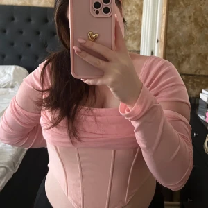 Rosa korsett-topp - Säljer en snygg rosa korsett-topp med långa ärmar och offshoulder-design. Toppen har en figurnära passform och är perfekt för en trendig look. Passar bra till både jeans och kjol.