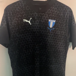 Malmö FF träningströja  - Säljer en svart fotbollströja från Puma med Malmö FF-logga på bröstet. Tröjan har ett diskret mönster och korta ärmar. Perfekt för fotbollsfans som vill visa sitt stöd för laget.
