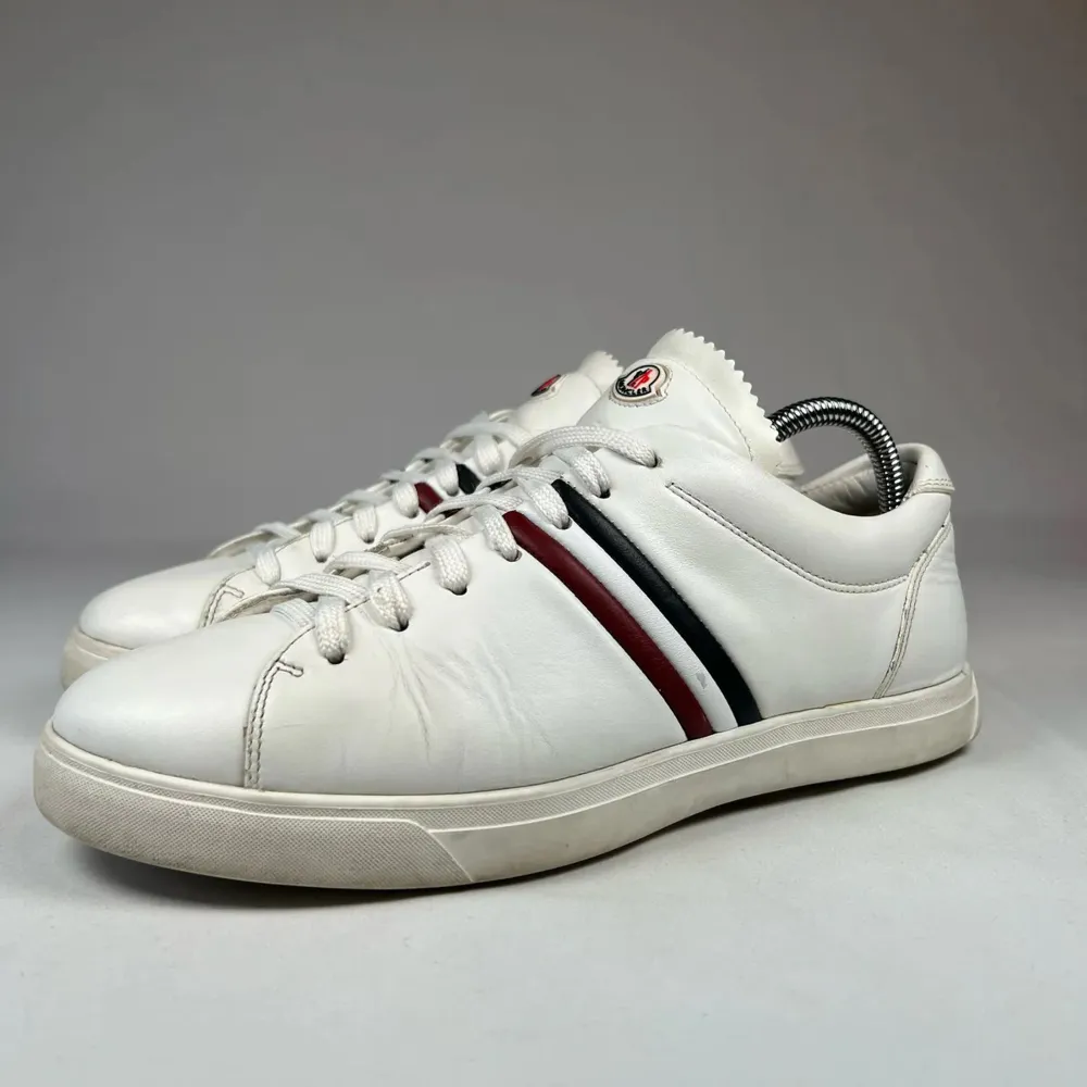 Snygga vita sneakers med klassiska ränder i svart och rött på sidan. Skorna har snörning och en rund tå, vilket ger en stilren look. Perfekta för en avslappnad stil.. Kengät.