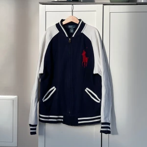 Blå och vit bomberjacka från Polo Ralph Lauren - Säljer en snygg zip-tröja / collegejacka från Polo Ralph Lauren i storlek L (14–16). Tröjan är i bra och fint skick och är inte använd så mycket.