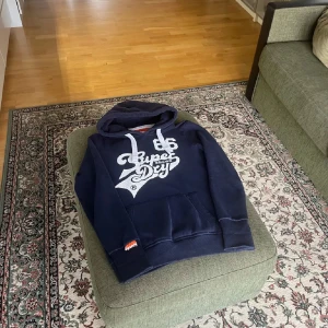 Mörkblå hoodie från Superdry - Säljer en snygg mörkblå hoodie från Superdry med stort vitt tryck på framsidan. Tröjan har en klassisk känguruficka och justerbar huva med vita snören. Perfekt för en avslappnad stil. 🧢
