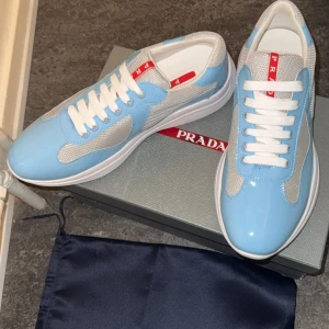 Blå och vita sneakers från Prada - Snygga sneakers från Prada i en fräsch ljusblå och vit färgkombination. Skorna har en stilren design med vita snören och en röd logga på plösen. Perfekta för en trendig look.