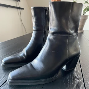 Svarta skinnstövletter från Vagabond - Snygga svarta stövletter i skinn från Vagabond med dragkedja på sidan. De har en fyrkantig tå och en stabil klack som ger en elegant look. Perfekta för att komplettera din outfit med stil.