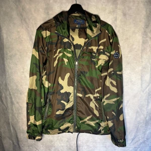 Camouflage vindjacka från Polo Ralph Lauren - Säljer en cool camouflage vindjacka från Polo Ralph Lauren. Jackan har en dragkedja framtill och en broderad logga på ärmen. Perfekt för en avslappnad stil med sin gröna och bruna färgkombination. Samt svarta Replay jeans samt Ralph Lauren tröja