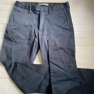 Mörkblå byxor från Acne Studios - Snygga mörkblå byxor från Acne Studios i modellen Max Satin. Tillverkade i en bomullsblandning med elastan för extra komfort. Byxorna har en klassisk design med dragkedja och knapp framtill samt fickor både fram och bak.