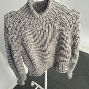 Grå stickad tröja - Säljer en stilren grå stickad tröja med hög krage och ribbade detaljer. Aldrig använd pågrund av fel storlek! Perfekt för kyliga dagar med sin mysiga och varma design. Tröjan har långa ärmar och en klassisk passform som passar till det mesta. Ett måste i garderoben för höst och vinter men även en kall sommar kväll!