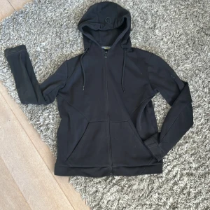 Svart hoodie med dragkedja - Säljer en stilren svart hoodie med dragkedja och huva. Den har långa ärmar och praktiska fickor framtill. Perfekt för en avslappnad look.