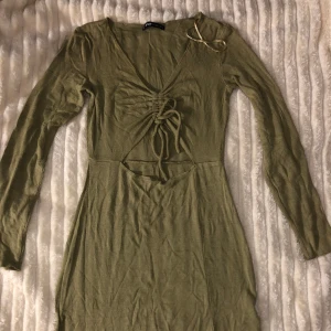 Grön ribbad klänning med knyt - Snygg grön ribbad klänning med lång ärm och v-ringning. Den har en knytning framtill som ger en fin detalj. Perfekt för en stilren look.