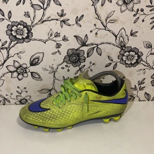 Gröna och lila fotbollsskor från Nike - Säljer ett par coola gröna fotbollsskor från Nike med lila detaljer och snörning. Skorna har en unik design med en dödskalle på sidan och är perfekta för gräsplaner. De har en mönstrad yta för bättre grepp och kontroll.