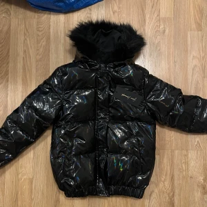 Svart glansig pufferjacka med huva - Snygg svart pufferjacka med glansig yta och fluffig huva. Perfekt för kyliga dagar. Jackan har långa ärmar och elastiska muddar för extra komfort💗💗