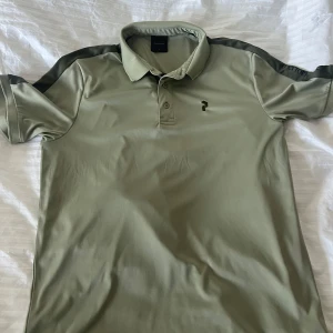 Olivgrön pikétröja från Peak Performance - Snygg olivgrön pikétröja från Peak Performance med korta ärmar och klassisk krage. Tröjan har en diskret logga på bröstet och text längs ärmen. Perfekt för en stilren och sportig look. Använd endast någon gång. Mycket bra skick