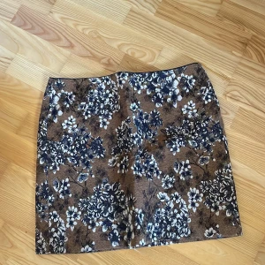 Blommig brun kjol - Säljer en snygg brun kortkjol med blommigt mönster i vitt och blått. Perfekt för att liva upp garderoben med lite mönster och färg. Kjolen har en normal passform och är lätt att matcha med olika toppar.