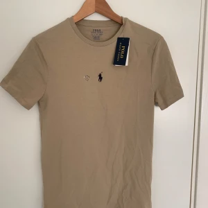 Beige t-shirt från Polo Ralph Lauren - Säljer en stilren beige t-shirt från Polo Ralph Lauren. Den har en klassisk passform med korta ärmar och är tillverkad i mjuk bomull. Den ikoniska loggan är broderad på bröstet. Perfekt för en avslappnad look.