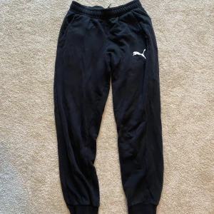 Svarta mjukisbyxor från Puma - Säljer ett par svarta mjukisbyxor från Puma med elastisk midja och snörning. Byxorna har en broderad logga på sidan och är perfekta för en avslappnad stil. De har ribbade muddar vid bensluten för en skön passform.