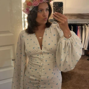 Adoore klänning - Charmig vit klänning med små blå blommor och en v-ringad design. Klänningen har långa ärmar och en slits framtill. Perfekt för sommarens festligheter. Pris kan diskuteras vid snabb affär