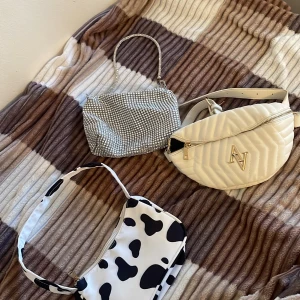 Tre trendiga väskor i olika stilar - Säljer tre snygga väskor: en glittrig silverväska med kedjehandtag, en vit quiltad magväska med dragkedja och en svartvit axelväska med ko-mönster. Perfekta accessoarer för att lyfta din outfit! 100kr för alla 3