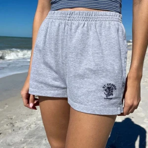 Grå shorts från Brandy Melville - Säljer ett par grå shorts från Brandy Melville i mjukt bomullsmaterial. De har en elastisk midja och en liten slits på sidorna för extra komfort. På framsidan finns ett broderat märke med texten 'St. George London'. Perfekta för en avslappnad dag eller träning!