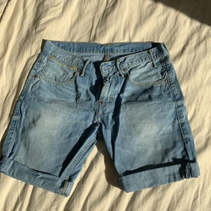 Blå jeansshorts från Levi's - Snygga blå jeansshorts från Levi's med klassisk femficksdesign och uppvikta ben. Perfekta för en avslappnad stil. Levi's logga syns tydligt på baksidan.
