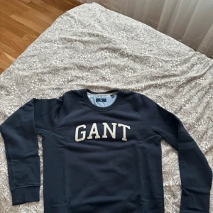 Mörkblå sweatshirt från GANT - Säljer en stilren mörkblå sweatshirt från GANT med deras logga i vitt på bröstet. Tröjan har långa ärmar och en rund halsringning. Perfekt för en avslappnad stil.