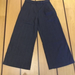 Grå, vida kostymbyxor Monki 34 - Gråmelerade kostymbyxor från Monki i storlek 34. Mjukt tyg, vida raka ben, pressveck upptill, fickor i sidorna, en fuskficka bak. 76% Polyester,23%  viskos, 1% elastane. Midja 33cm, midjehöjd 26cm, höft 52 cm, övre lår 32 cm, ben 29cm, innerbenslängd 60cm