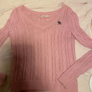 Rosa stickad tröja från Abercrombie & Fitch - Säljer en söt rosa stickad tröja från Abercrombie & Fitch med kabelstickat mönster och en liten logga på bröstet. Tröjan har en v-ringad hals och långa ärmar. Perfekt för kyliga dagar!